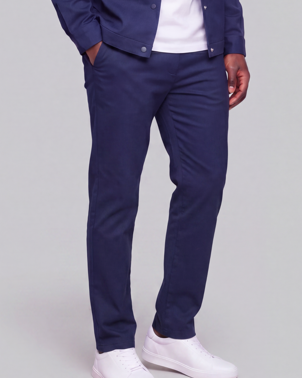 A person wearing navy blue slim-fit trousers, styled with a white t-shirt, a navy jacket, and white sneakers, shown from the waist down on a light gray studio background.