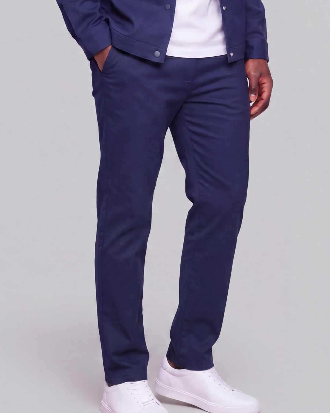 A person wearing navy blue slim-fit trousers, styled with a white t-shirt, a navy jacket, and white sneakers, shown from the waist down on a light gray studio background.